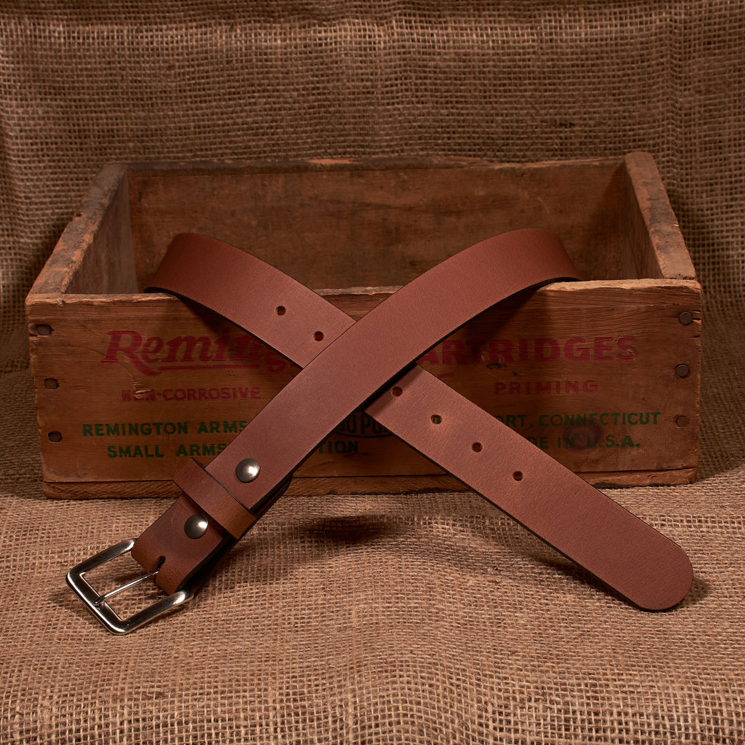 Brown leather belt with silver buckle on a wooden crate labeled 'Remington Arms' against a burlap background.