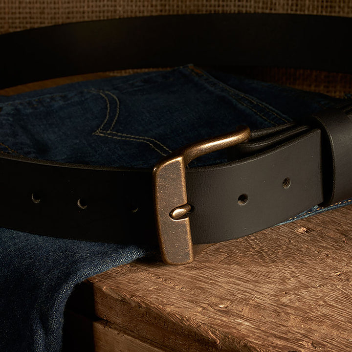 Dark leather belt with brass buckle on a pair of blue jeans on a wooden surface.