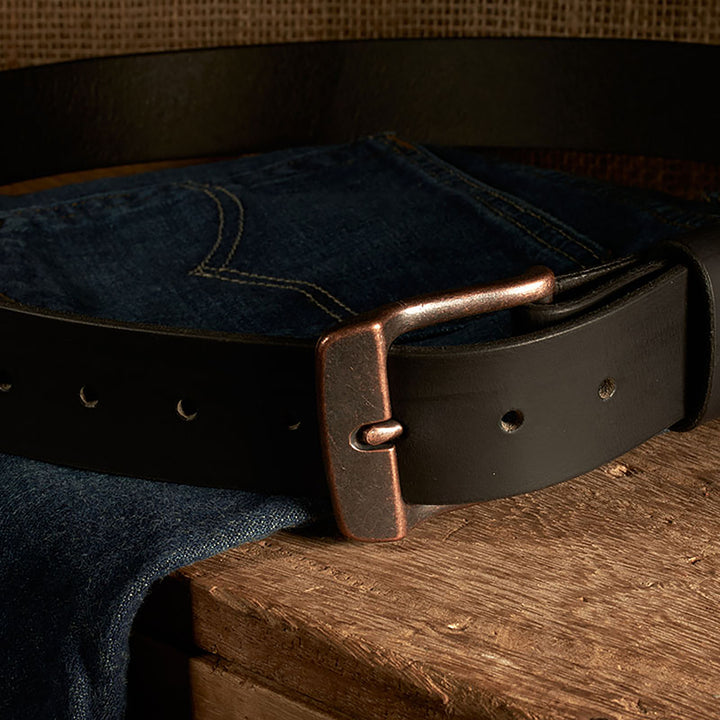 Dark leather belt with a bronze buckle on a pair of blue jeans on a wooden surface.