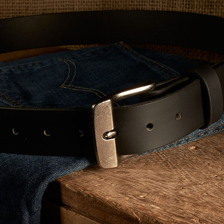 Black leather belt with brass buckle on a pair of blue jeans on a wooden surface.