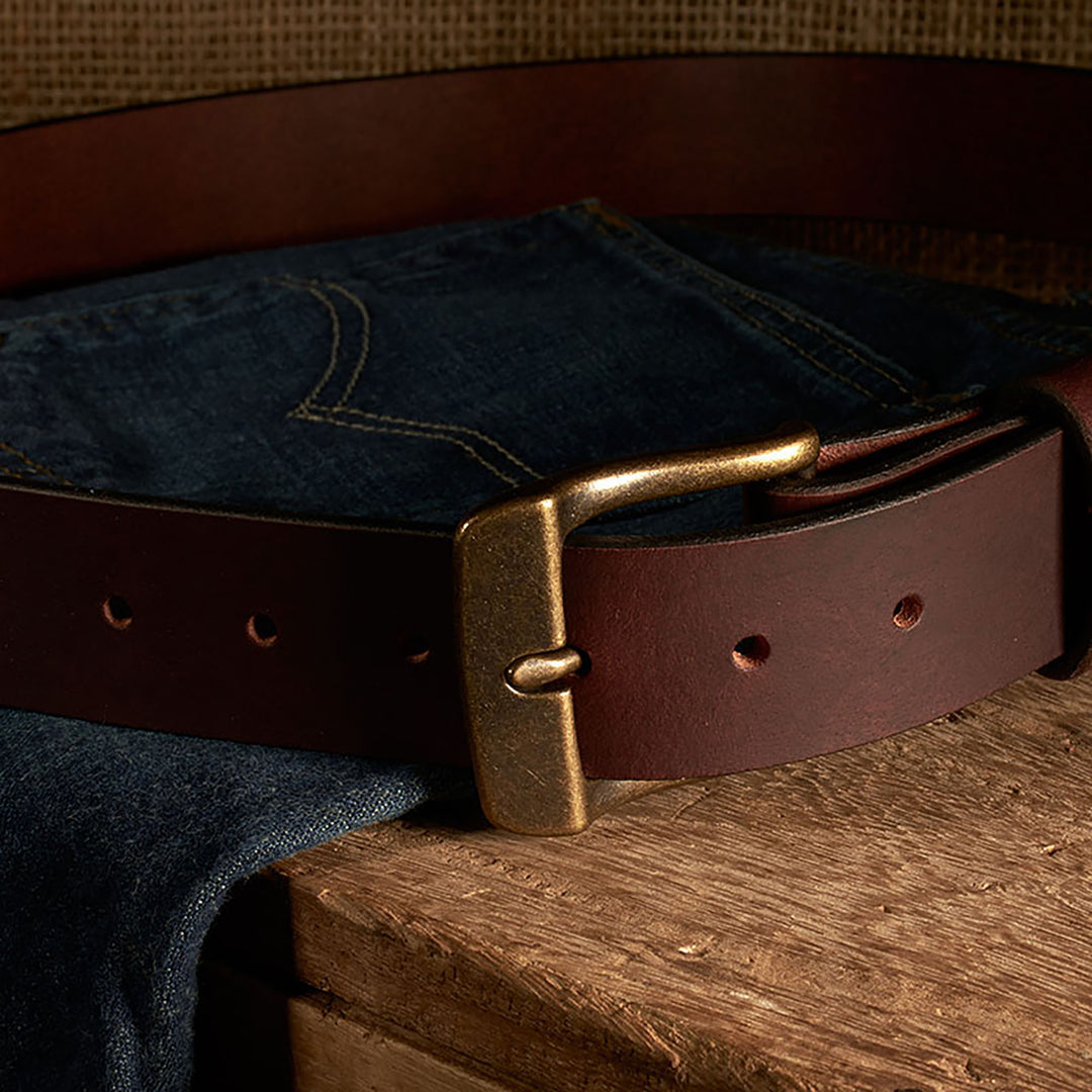 Brown leather belt with brass buckle on a pair of blue jeans on a wooden surface.