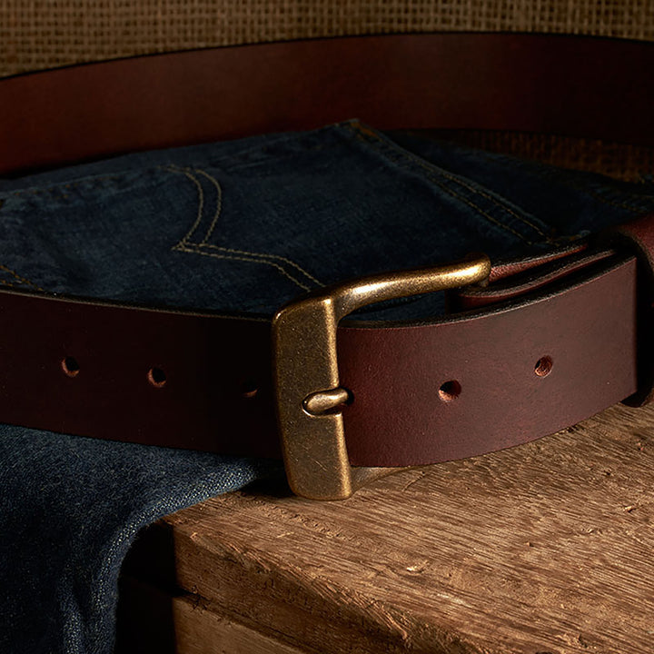 Brown leather belt with brass buckle on a pair of blue jeans on a wooden surface.