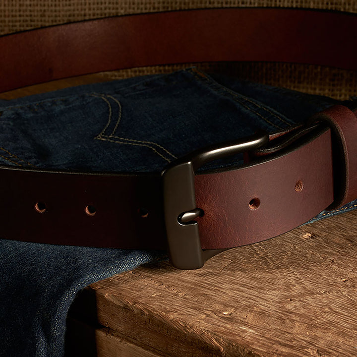 Brown leather belt with a metal buckle on blue jeans against a wooden surface.
