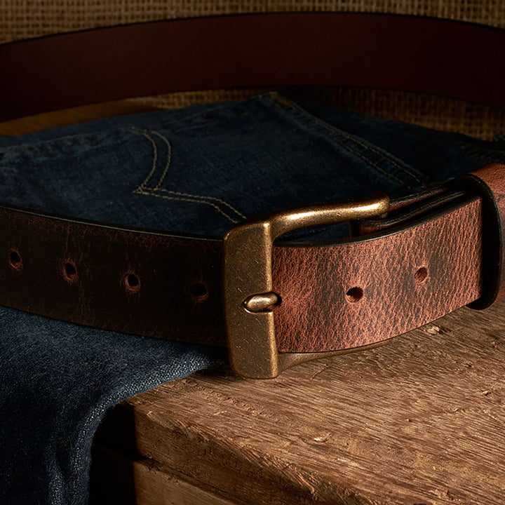 Brown leather belt with brass buckle on a pair of blue jeans on a wooden surface.