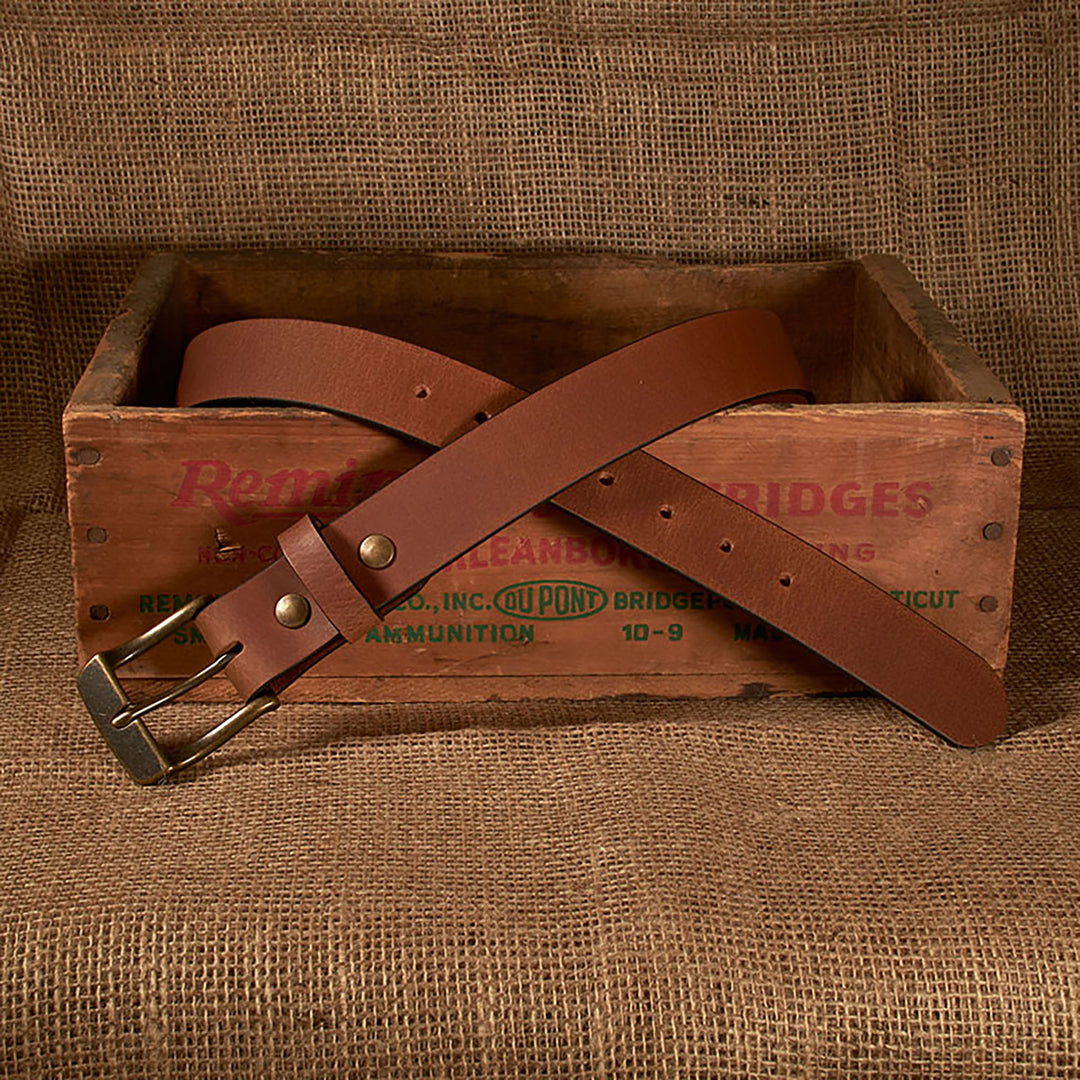 Brown leather belt with brass buckle on a wooden box with text, against a brown fabric background.
