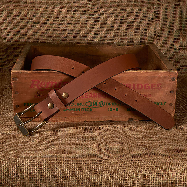 Brown leather belt with brass buckle on a wooden box with text, against a brown fabric background.