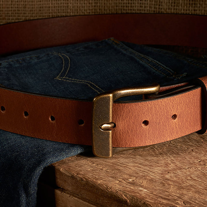 Brown leather belt with brass buckle on a pair of blue jeans against a wooden surface.