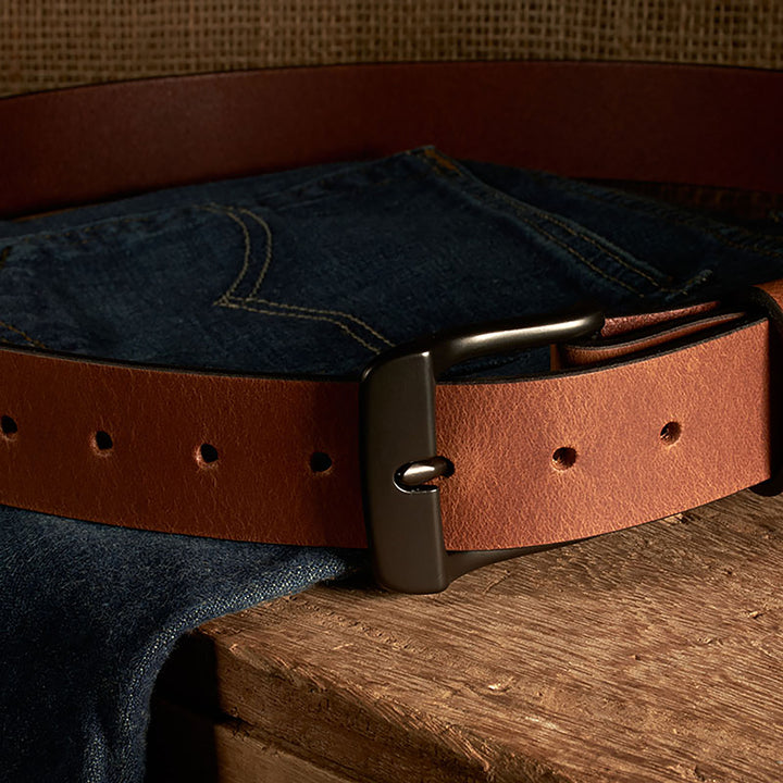 Brown leather belt with a metal buckle on a pair of blue jeans against a wooden surface.