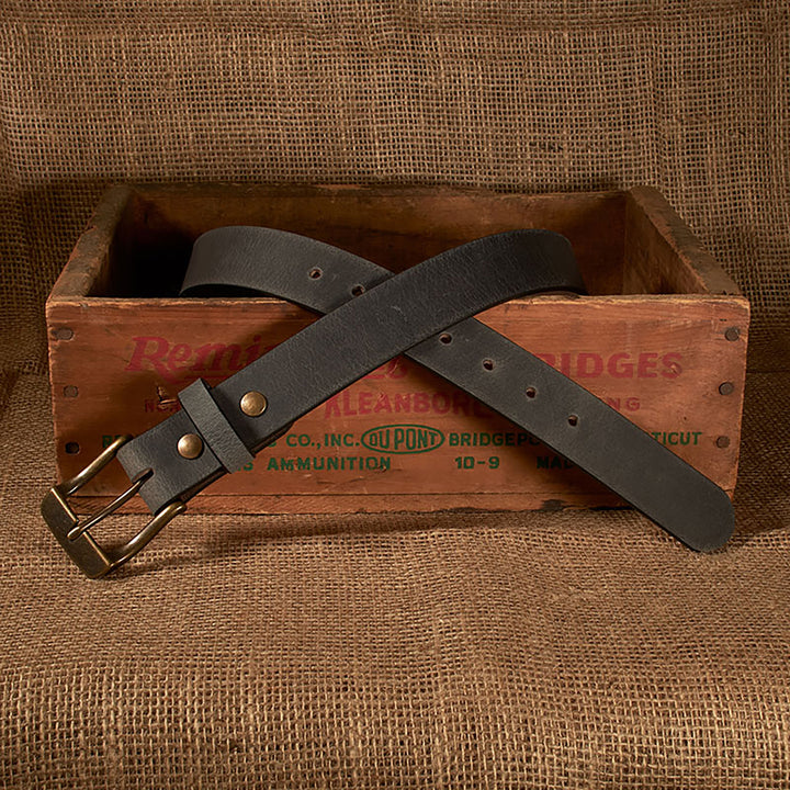 Dark leather belt with brass buckle on a rustic wooden box against a brown fabric background