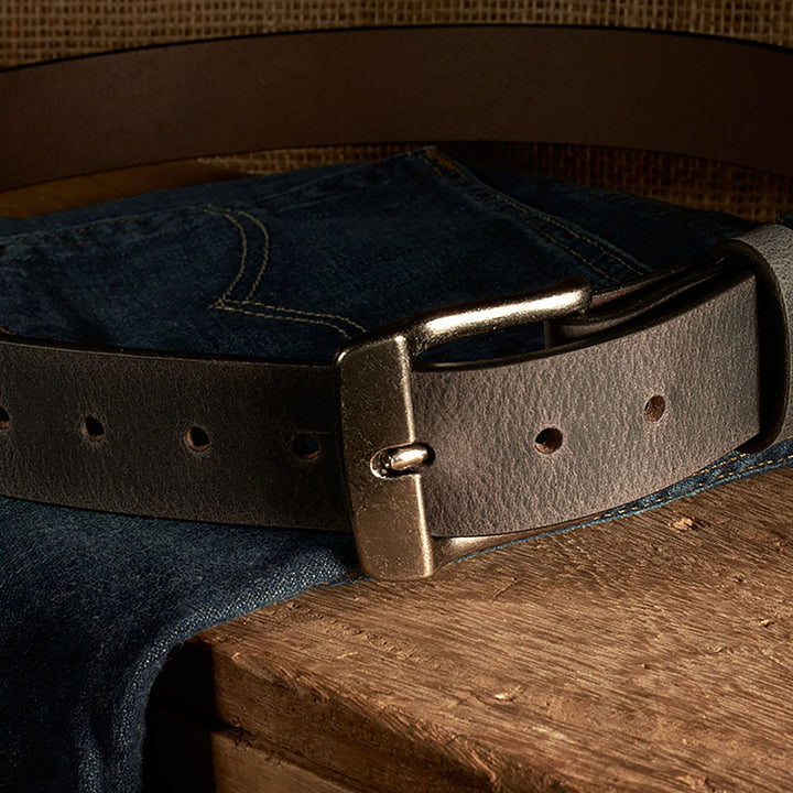 Brown leather belt with brass buckle on a pair of blue jeans on a wooden surface.
